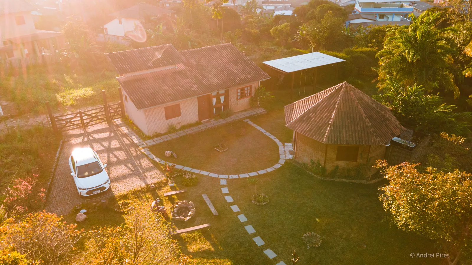 Vista aérea da hospedagem ao pôr do sol, com a casa principal, a cabana redonda, o jardim e a fogueira