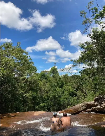 Casal sentado na correnteza no topo da cachoeira, de frente para a mata e o céu azul