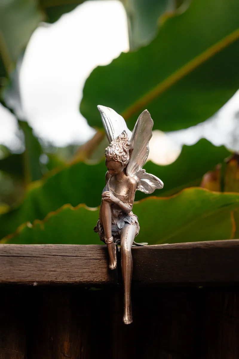 Estatueta de fada de bronze sentada em uma viga de madeira, entre as folhas verdes do jardim