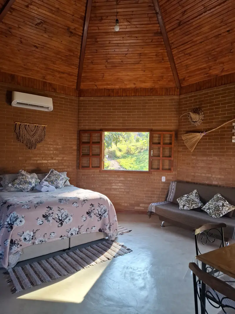 Interior de cabana com teto de madeira em formato octogonal, cama de casal e janela aberta para o verde