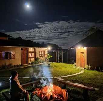Quintal da Casa do Chapéu à noite, com fogueira acesa, lua cheia e as duas cabanas iluminadas
